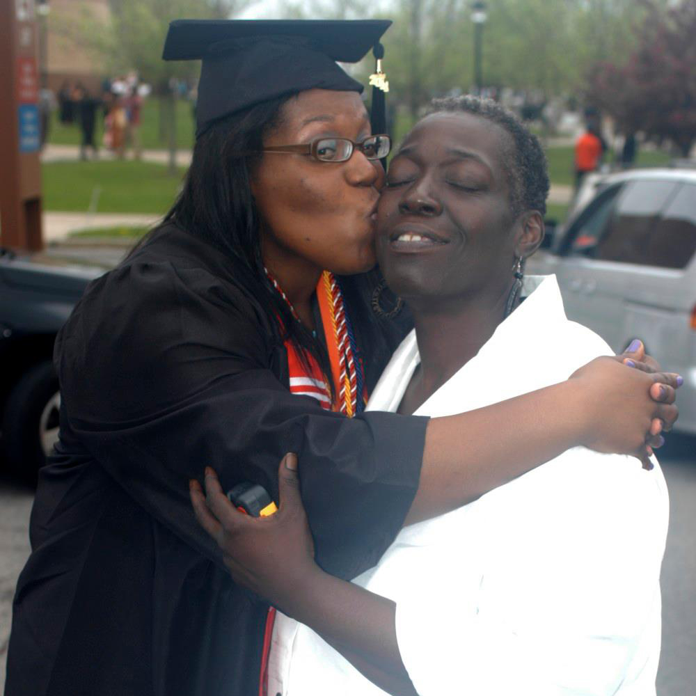 Two women, one dressed in graduation clothing and kissing the cheek of the other woman. 